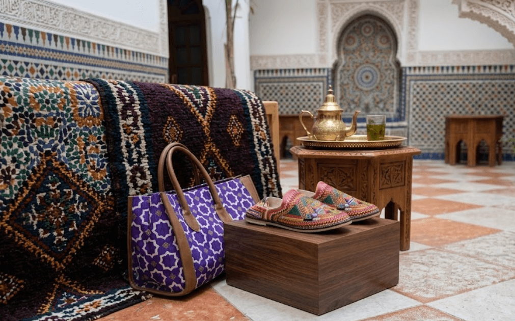 Handmade embroidered Moroccan babouche leather slippers displayed on a wooden stand in a traditional riad courtyard with zellige tilework, a brass Moroccan teapot, and a mint tea glass in the background